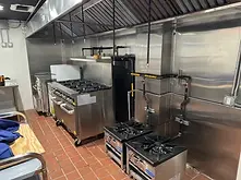 Commercial kitchen with stainless steel appliances, including a range and two smaller burners, under a large vent hood with exposed metal pipes, labeled New Construction below the image.