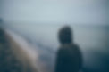 Betrayal Trauma. Image shows the back of a person wearing a hood, standing alone on a foggy beach or shore, suggesting isolation and contemplation of loss.