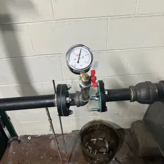Close-up of a pressure gauge and a red-handled shut-off valve installed on a black metal gas line, secured with black and red flanges, against a white cinder block wall.