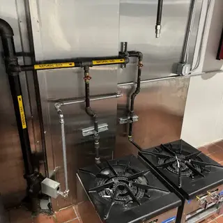 Full view of a commercial kitchen setup with stainless steel range, two countertop burners, and an exhaust hood system with black gas lines and safety equipment visible.