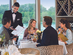 River cruise ship dining room set for an elegant dinner, white linen tablecloths, wine glasses, warm candlelight, passengers dressed for an evening meal
