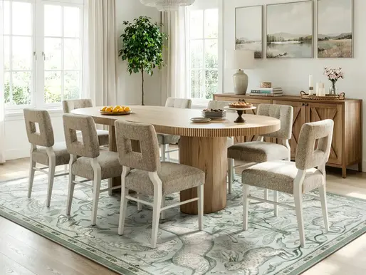 Bright modern dining room with oval wood table, six beige chairs, fruit and desserts, large windows, plants, and patterned rug.