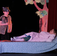 Children performing a theatre scene on stage with props and a colourful set.