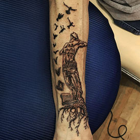 Black and grey human figure tattoo with birds on the forearm.