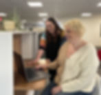 Two employees collaborate in a bright office space, with one seated at a desk typing on her laptop and showing her screen to a smiling coworker standing nearby.