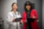 Two Ohio State University Physicians Community Medical Group doctors, affiliated with The Ohio State University Wexner Medical Center, stand in front of a patient room door at an outpatient care location, holding clipboards and smiling. One wears a white coat with a stethoscope, and the other a red blazer with a colorful stethoscope.