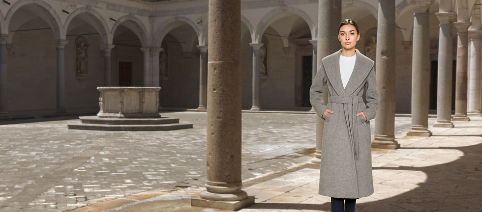 Model wearing a belted light-grey coat over a white top and dark trousers in the stone courtyard of Montecassino Abbey, Italy.