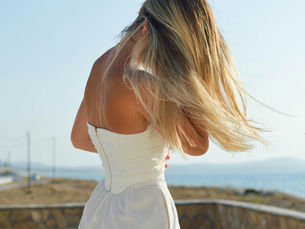 Woman in a white dress standing on a rocky cliffside during daytime, looking out at the ocean with wind blowing through her long hair.