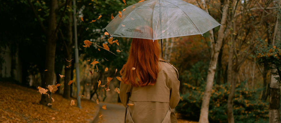 Woman with red hair in a trench coat walks on an autumn path holding a clear umbrella.
