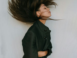 Woman in a black coat flipping her long hair while looking confidently into the distance against a neutral background.