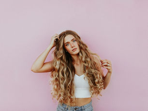 Woman with long wavy hair standing in front of a textured pink wall, casually scratching her head and looking relaxed and stylish in a neutral-toned outfit.