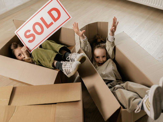 Two young children play inside moving boxes on the floor of their new home. One child holds a bright red “SOLD” sign while the other raises their hands with excitement, capturing the joy of moving into a new house.