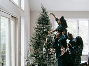 A family decorating a Christmas tree together in a bright living room, with an adult lifting a child to place a star on top, representing home, stability, and the holiday season.