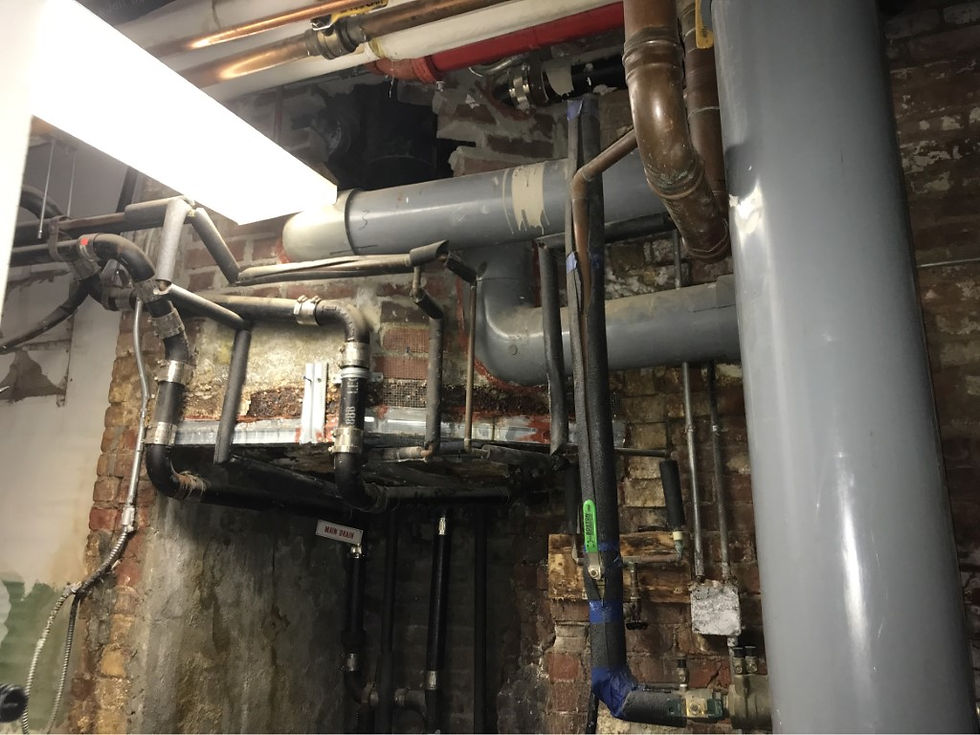 Complex network of pipes against a brick wall. Rust and corrosion visible, with a "Main Drain" sign. Industrial setting.