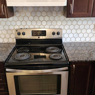 Installation of white hexagon tile backsplash with gold trim in a kitchen with brown cabinets.