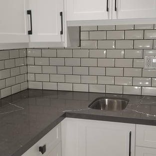 Installation of white subway tile backsplash in a kitchen with white cabinets, and grey countertops.