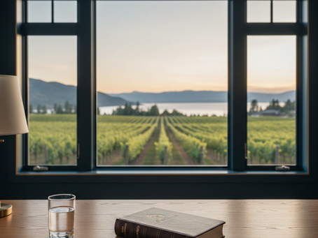 Glass of water and book on a wooden table by a window with a vineyard view, during sunset. Calm ambiance with mountains and lake outside.