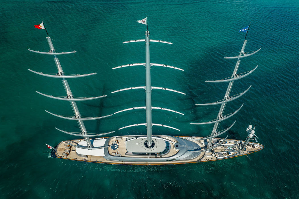 Aerial view of a large sailing superyacht with fully raised sails over calm blue water.