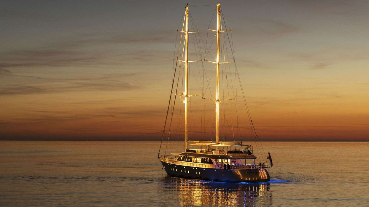 Aurum Sky sailing yacht anchored offshore with masts lit during a sunset charter