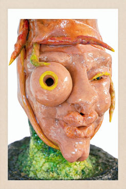 Detailansicht einer Büste mit orangefarbenen, wurmartigen Formen auf dem Kopf, einem seitlich hervortretenden runden Auge mit gelber Öffnung, grünem Hals und dunkel gepunktetem Sockel.