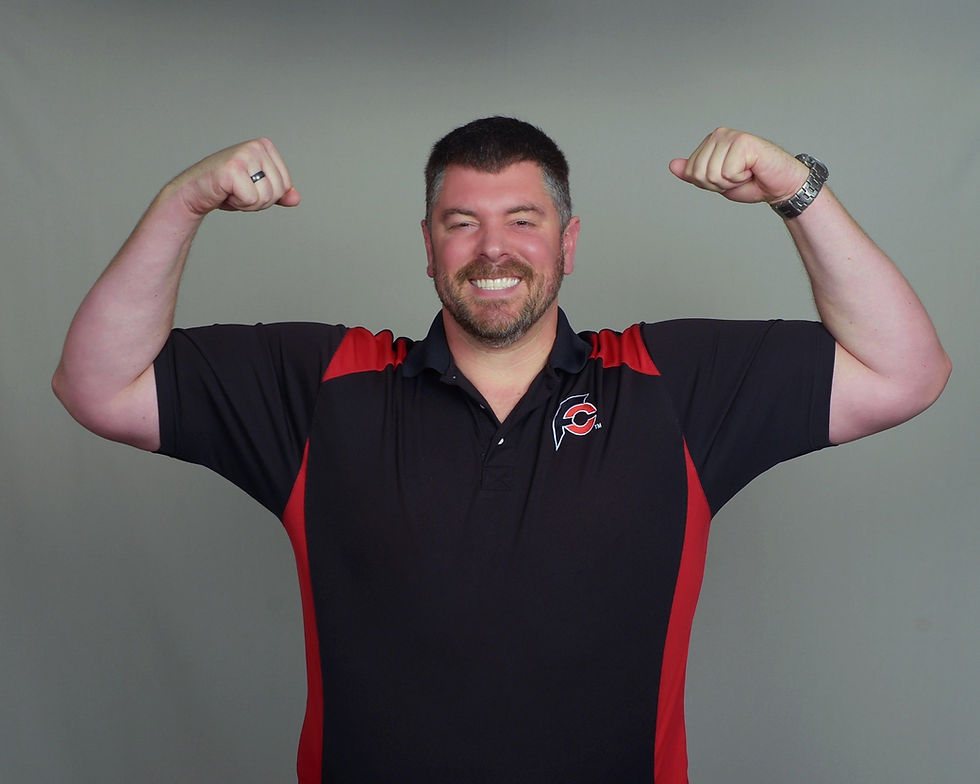 Randy Santel (Atlas) in black and red shirt flexes arms, smiling broadly. Gray background. Shirt has a small logo with a letter. foodchallenges.com Positive mood.