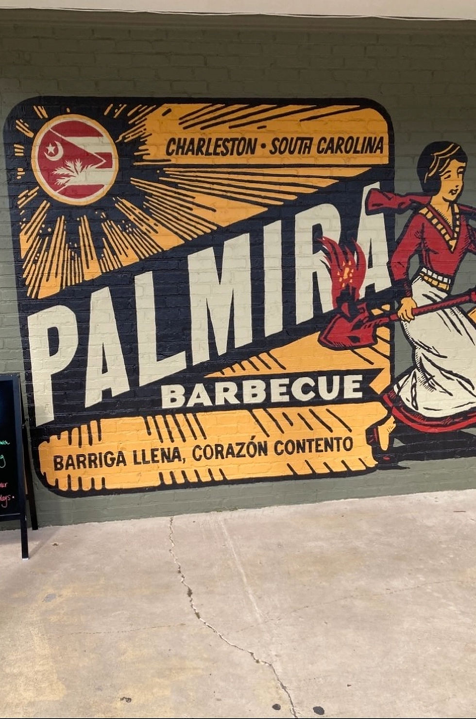 Colorful mural for Charleston, South Carolina Palmira Barbecue with a woman carrying a shovel and Spanish text, Barriga llena, corazón contento