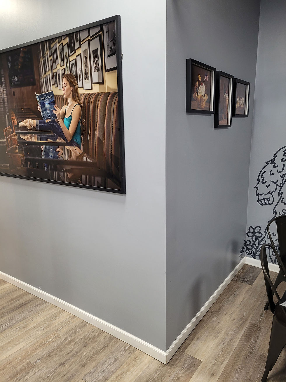 Framed photo of a woman reading on a cafe wall. Gray walls with additional small framed pictures. Wood floor, cozy atmosphere.