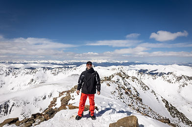 Hiker standing on a snowy Colorado 14er summit with panoramic Rocky Mountain views.