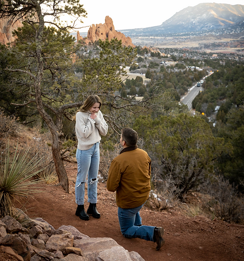 Surprise proposal at Garden of the Gods in Colorado Springs with man kneeling on a scenic overlook surrounded by pine trees and mountain views