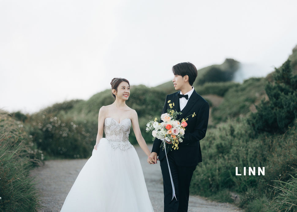 	山路牽手婚紗，新娘深情看著新郎。 / Couple holding hands on mountain road, bride gazing at groom.