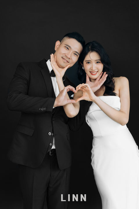 黑色背景趣味婚紗，新人互相捧臉並一起比愛心。 / Playful couple holding each other's faces and making a heart shape.