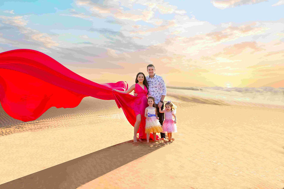 Family of four in desert at sunset. Woman in flowing red dress, man in floral shirt. Two girls in colorful dresses. Bright sky.