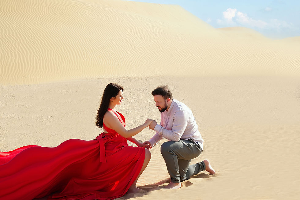 A romantic moment in a desert landscape with two individuals, one in a red dress seated on sand and the other kneeling and holding hands, under a clear sky with clouds.
