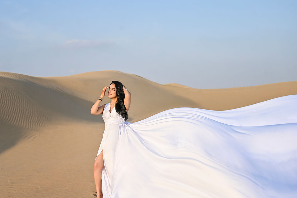 A woman in a flowing white dress stands in a desert landscape, with sand dunes in the background. She poses gracefully, with one hand in her hair and a serene expression, as the fabric of her dress flows dramatically behind her in the wind. The sky is clear and blue, enhancing the tranquil atmosphere of the scene.