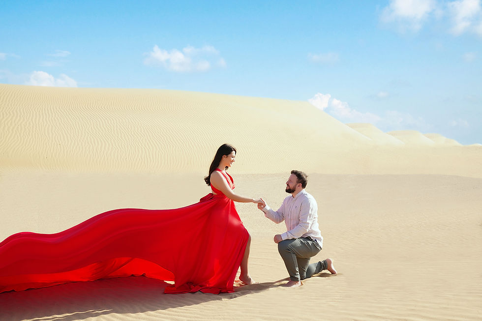 A couple in a desert setting, with the woman in a red dress and the man proposing on one knee, under a clear blue sky with sand dunes in the background.