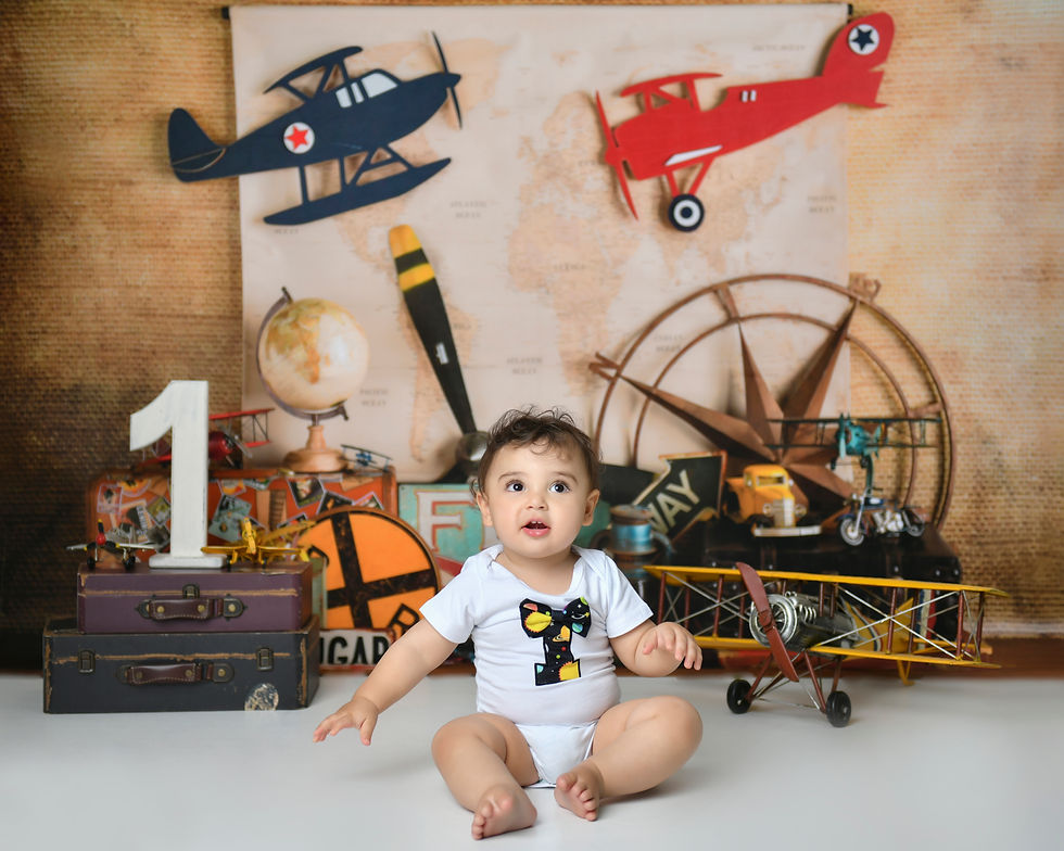 The image shows a baby sitting on a cake smash setup with a vintage aviation theme. The baby is wearing a white onesie with a bow tie and is surrounded by various vintage airplane models, globes, and other travel-themed props. The background features a map and illustrations of airplanes, creating a whimsical and adventurous setting for the baby's first birthday celebration.