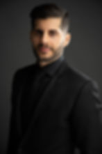 A man in a black suit with a tie, styled dark hair and beard, standing against a gray background with a neutral expression