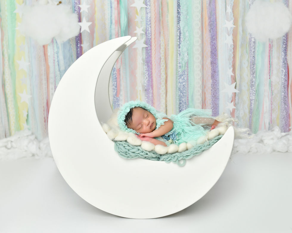The image shows a newborn baby sleeping peacefully in a crescent moon-shaped prop or cradle. The baby is wrapped in a soft, mint-colored blanket and wearing a matching headband. The background is a colorful, pastel-colored backdrop with stripes and stars, creating a whimsical and dreamy setting for the newborn portrait.