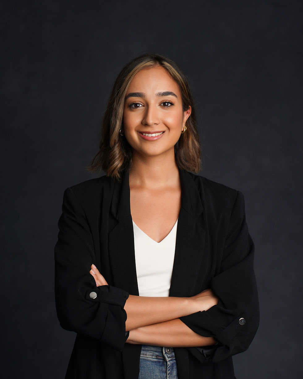 The image shows a professional headshot of a young woman with long brown hair and a warm, friendly smile. She is wearing a black blazer and a white shirt, and is standing in front of a dark background. The image conveys a sense of professionalism and confidence.