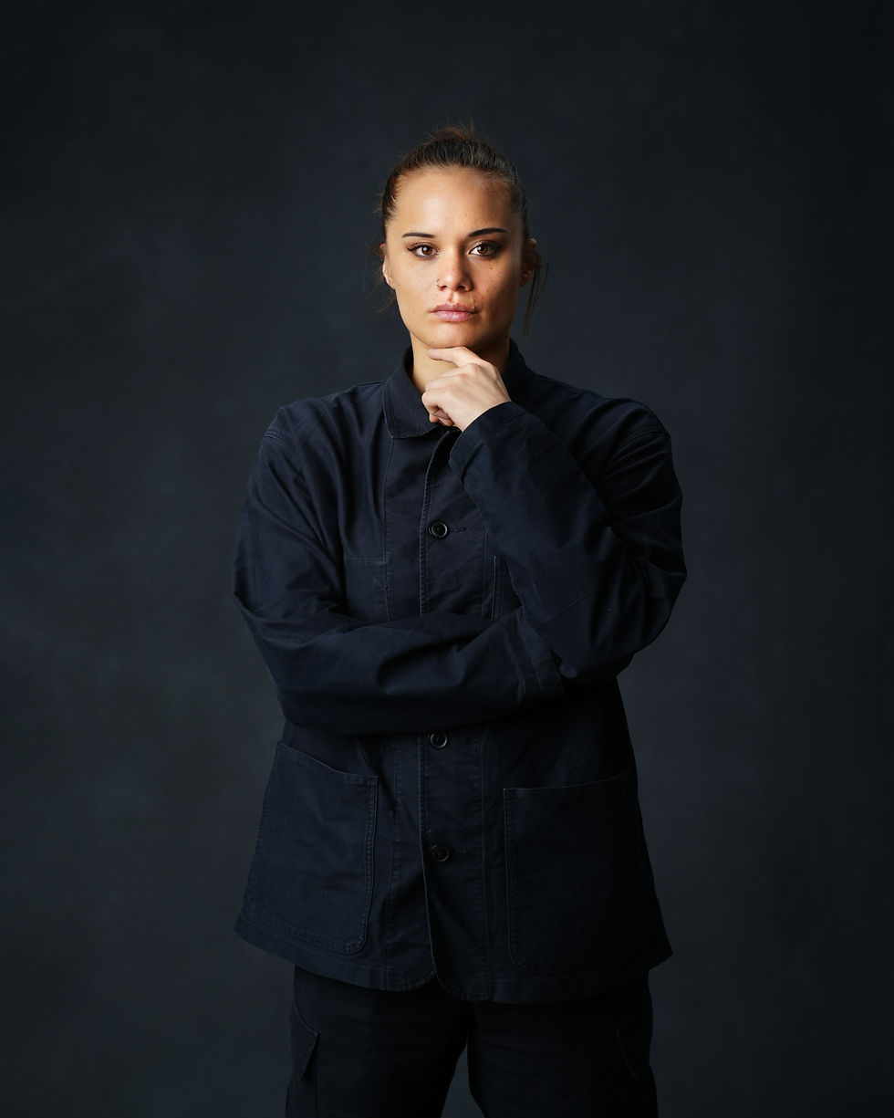 The image shows a young woman with dark hair wearing a black jacket, standing in a dark setting with a serious expression on her face. She has her hand on her chin, appearing to be deep in thought.