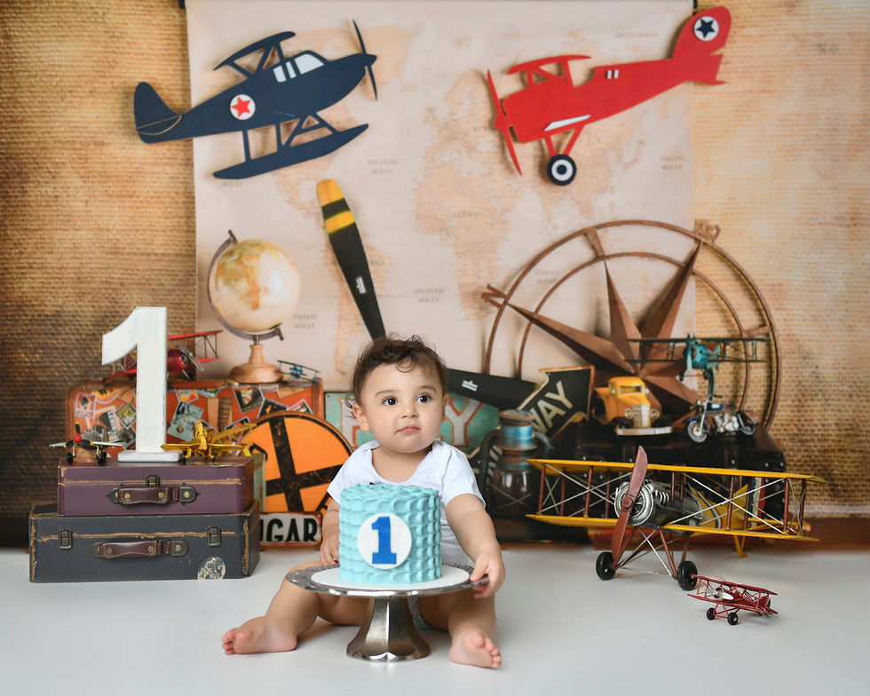 The image shows a young baby sitting on a cake on a pedestal, surrounded by various vintage airplane and travel-themed props and decorations. The background features a map and illustrations of airplanes, creating a whimsical and adventurous setting for the baby's first birthday cake smash photoshoot.