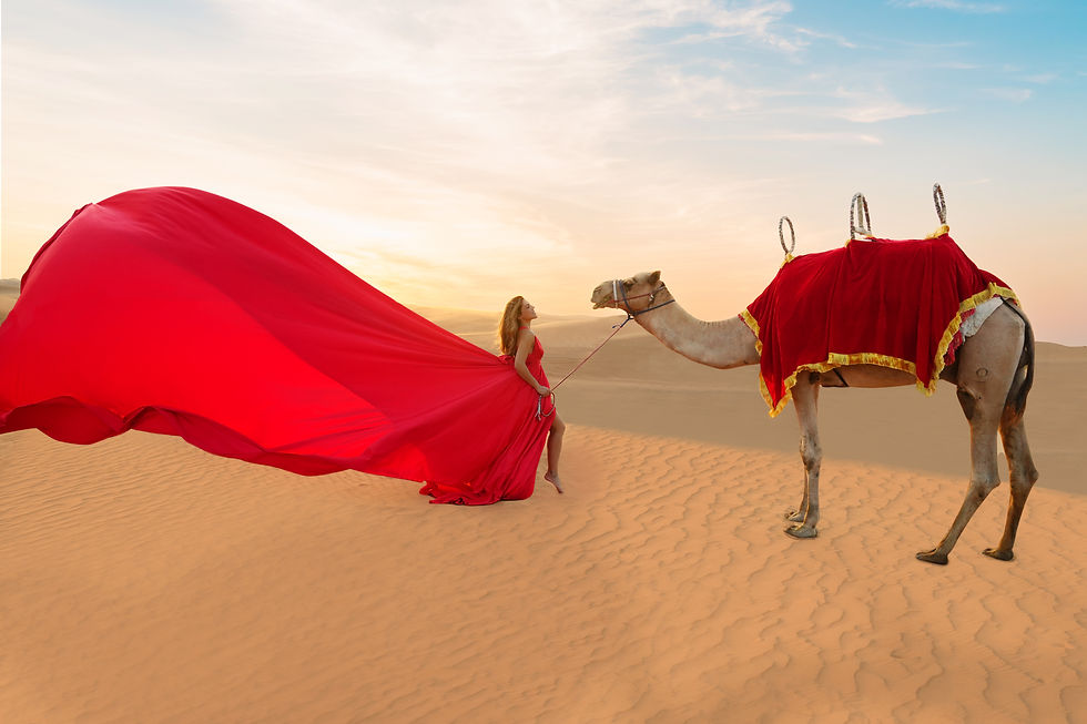 The image shows a woman in a flowing red dress standing in the desert, with a camel in the foreground also wearing a red ornamental covering. The sky is filled with warm, golden hues, creating a dramatic and picturesque desert scene.