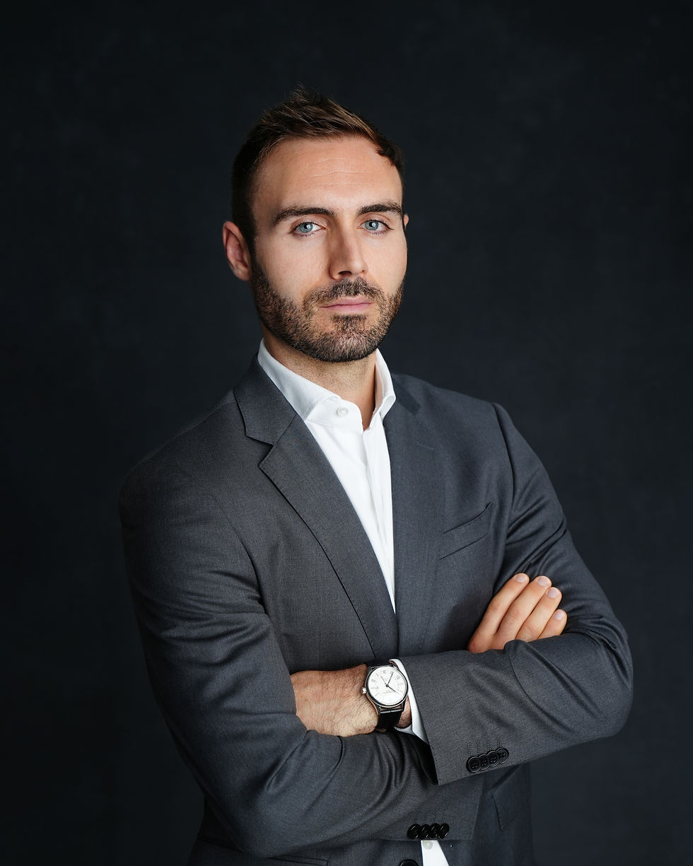The image shows a professional headshot of a young man with dark hair and a beard, wearing a gray suit and white shirt. He has a serious expression on his face and is standing in front of a dark background, conveying a sense of professionalism and confidence.