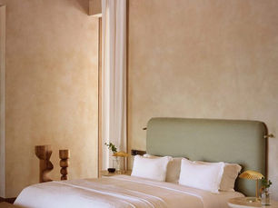 Cozy bedroom with a green upholstered bed, white linens, and wooden accents. Beige walls, woven ceiling, and soft natural light evoke warmth.