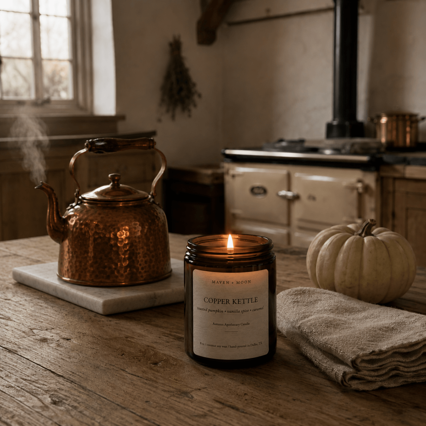 Lit Copper Kettle fall candle beside a hammered copper kettle and white pumpkin in a warm rustic kitchen.