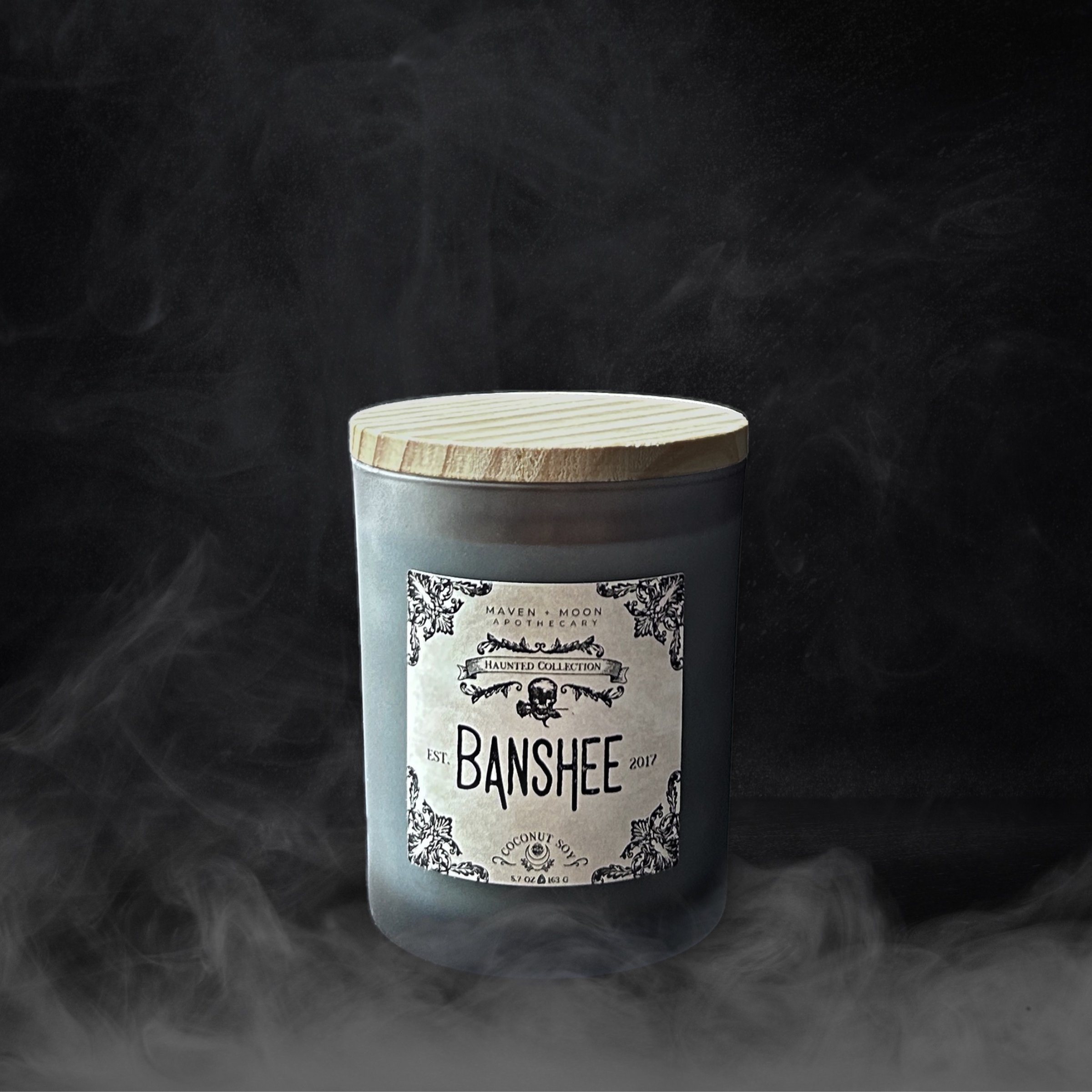 A grey blue clear frosted candle tumbler, bamboo lid, and vintage gothic label sit on a black background with billowing smoke