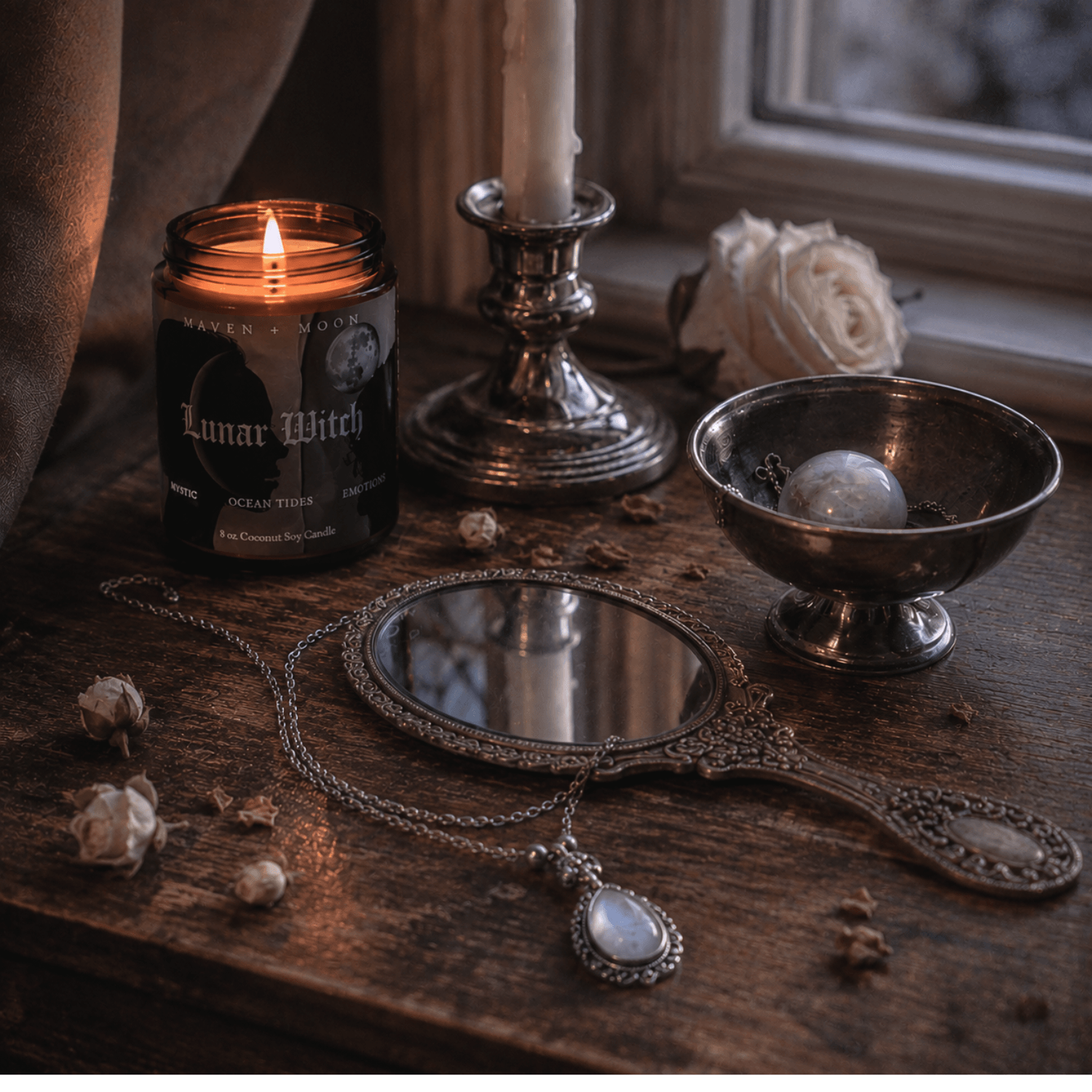 Lit Lunar Witch candle on a wooden windowsill beside an antique silver hand mirror and candle holder, illuminated by soft eve