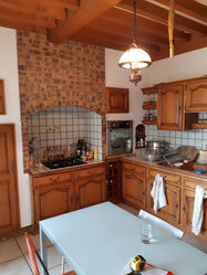 Cuisine rustique avant rénovation : meubles en bois massif, hotte en briques, carrelage mural à motifs, et plafond à poutres apparentes dans une maison familiale.