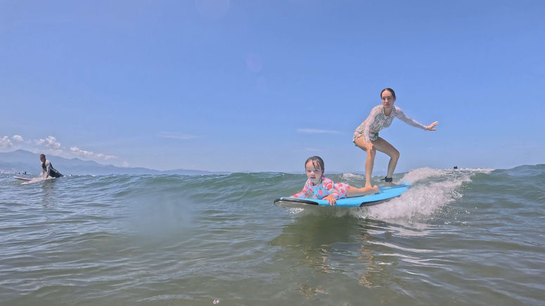 Mother and daughter surfing together on the same board during a family surf lesson with Surfing With Rudy Vallarta in Puerto Vallarta, Mexico.