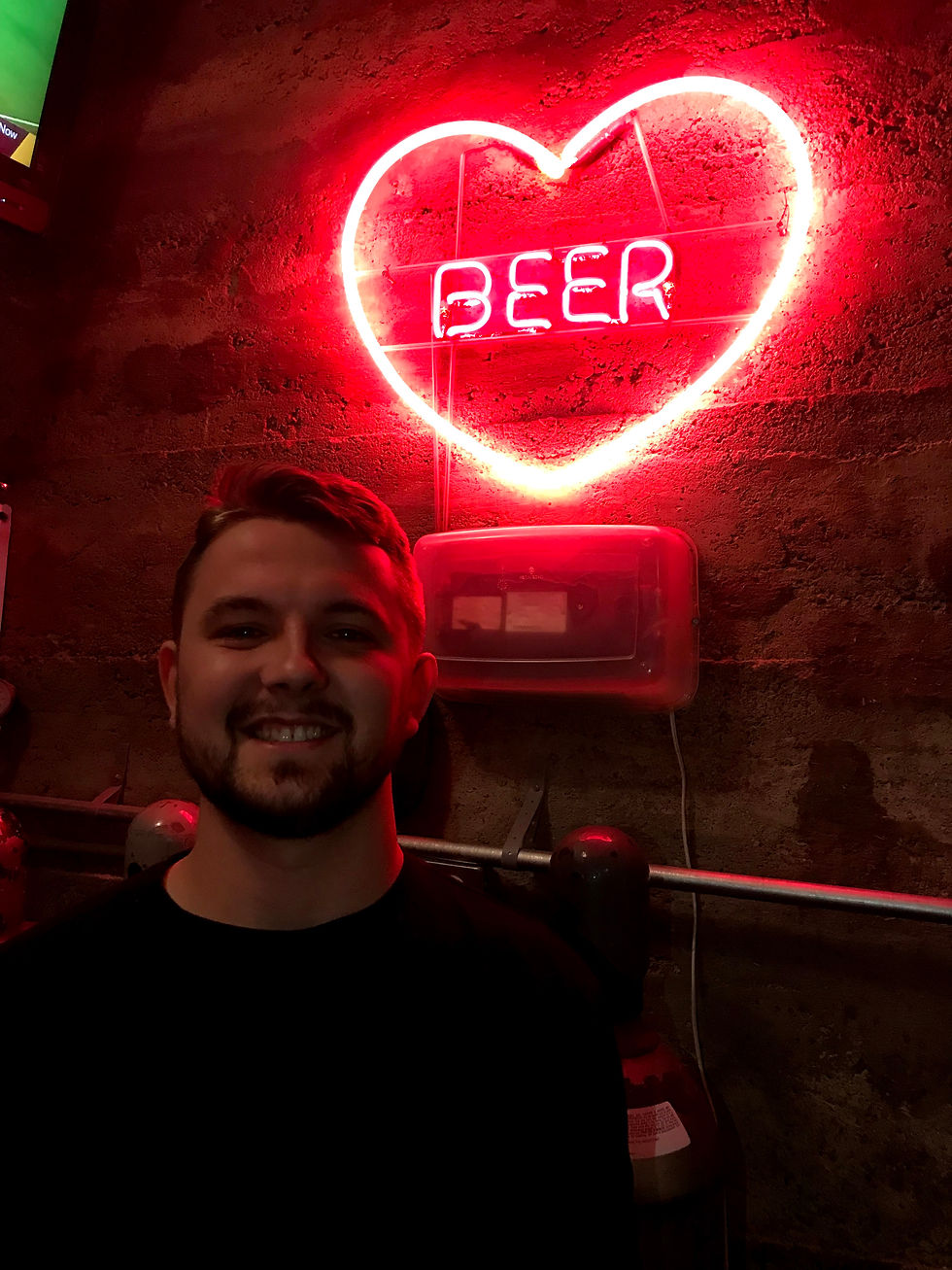 man smiling in front of neon heart sign that says, "BEER"
